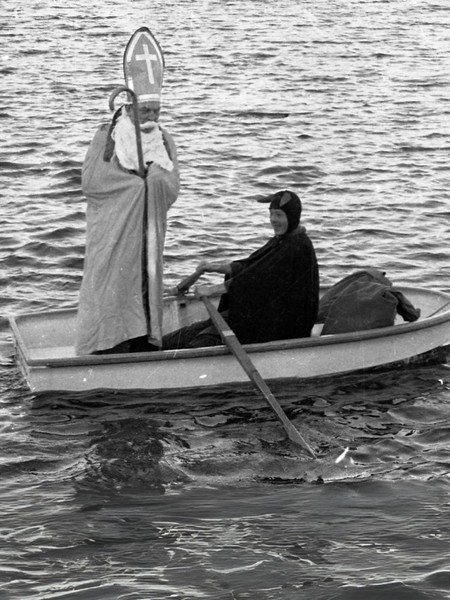 Der Nikolaus steht auf einem Boot auf dem Heiligen Meer. Ein schwarz gekleideter Mann rudert.